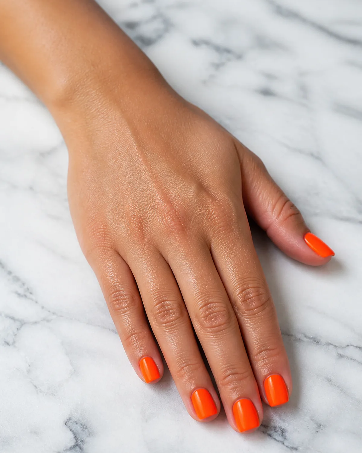 Single hand with electric neon orange gel on short square nails resting flat on white marble countertop in bright natural light