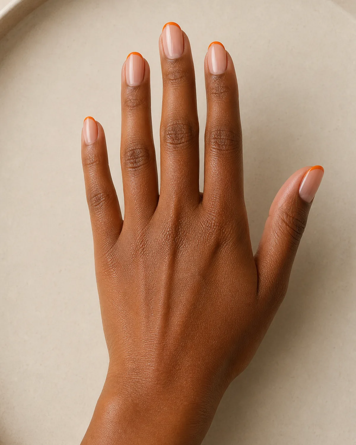 Single hand with sheer nude base and thin orange creme tips on medium oval nails resting palm-down on pale beige ceramic tray