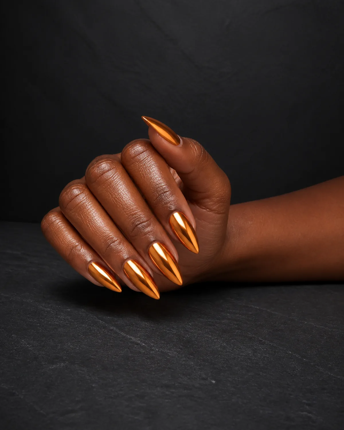 Single hand with mirror chrome finish in warm orange-copper on long stiletto nails resting on dark charcoal slate surface