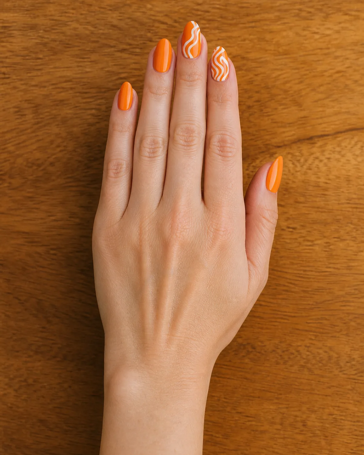 Single hand with glossy orange base and white swirl accents on two almond nails resting palm-down on warm wood surface