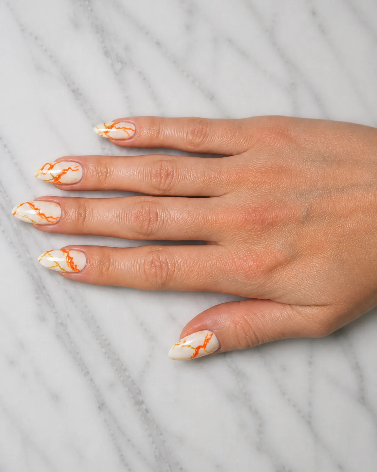 Single hand with white base and fine orange and gold veined marble pattern on medium almond nails resting palm-down on white marble countertop