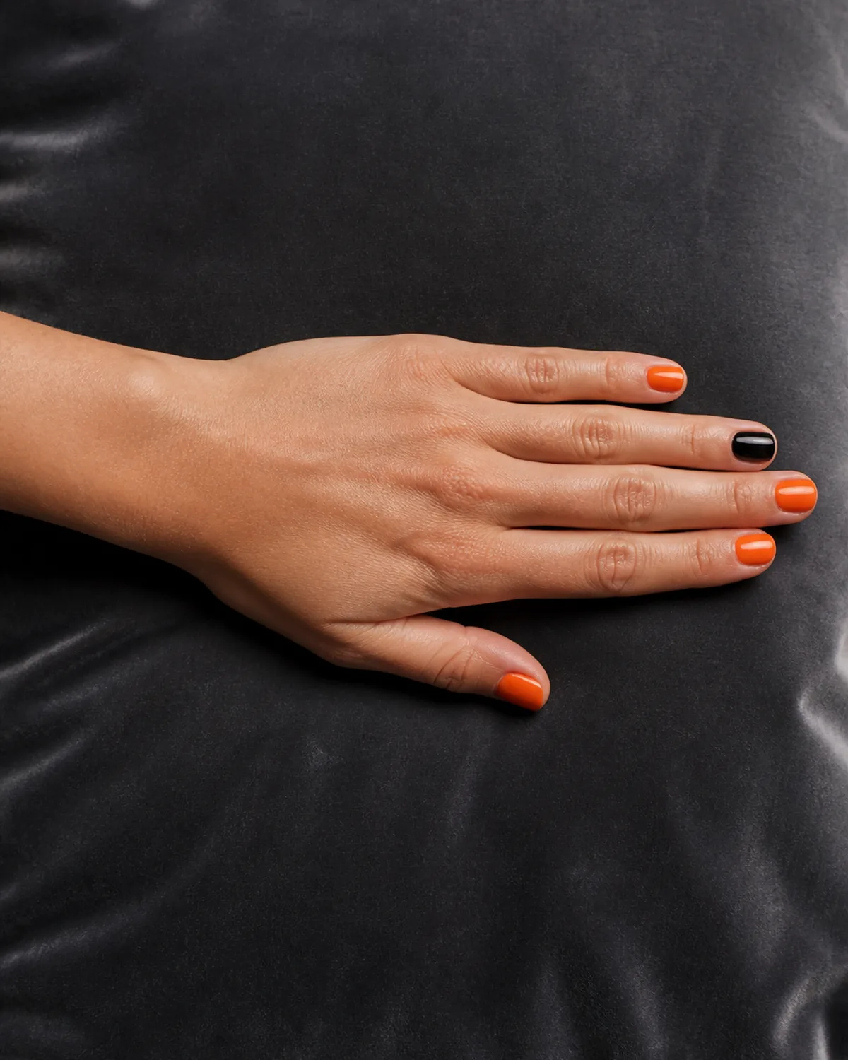 Single hand with glossy orange polish on round nails with one accent nail in solid black resting on dark gray velvet cushion