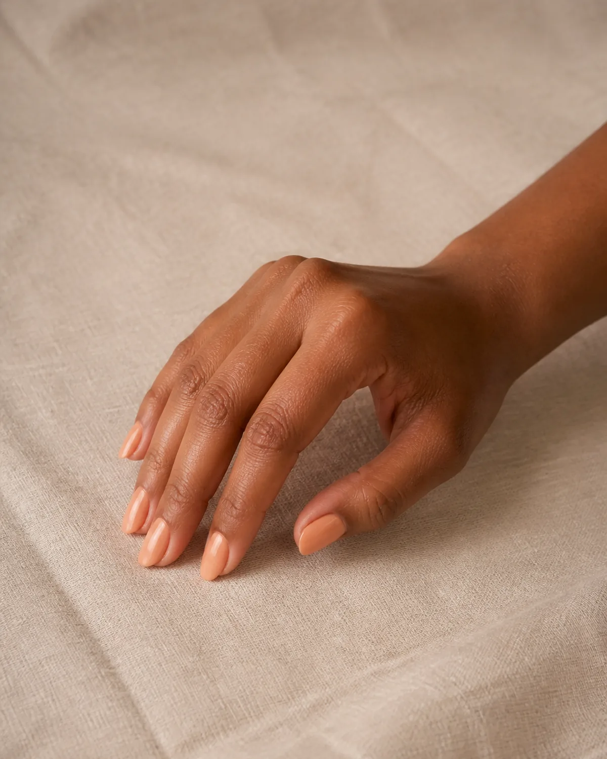 Single hand with muted warm apricot creme polish on medium oval nails resting at an angle with fingertips on warm beige linen surface