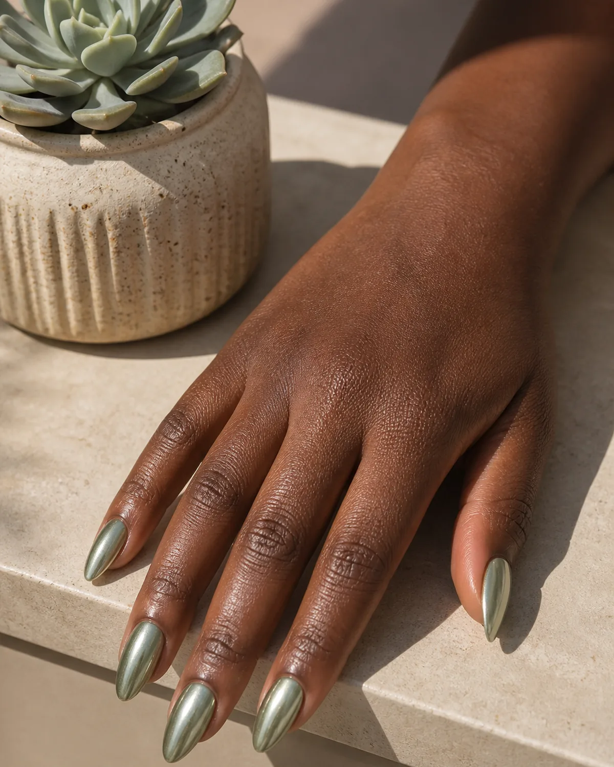 Single hand with sage-tinted chrome almond nails showing metallic green sheen beside a potted succulent on a beige shelf with dark skin tone