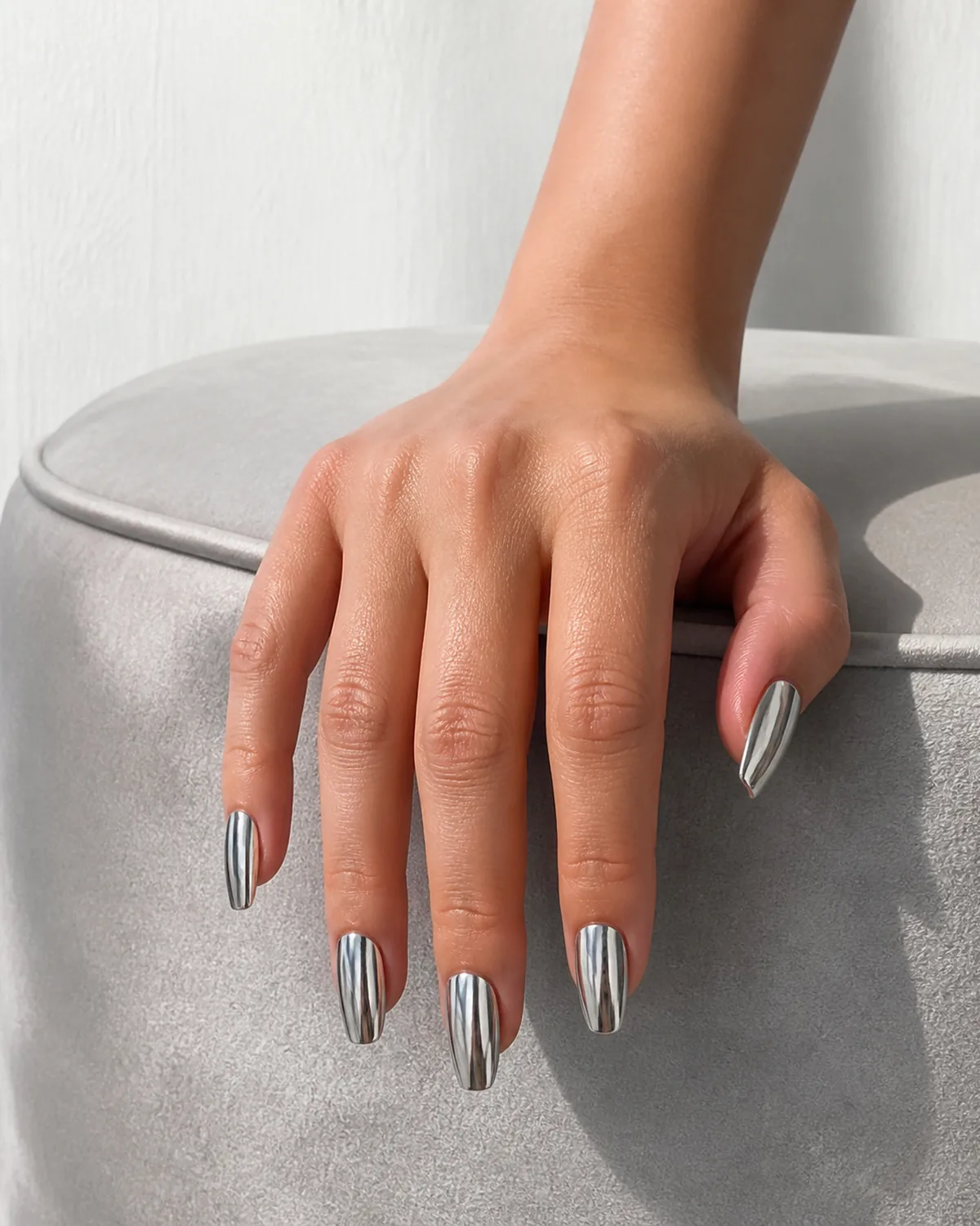 Single hand with opaque silver metallic polish with smooth reflective liquid-metal finish on medium coffin nails draped over pale gray velvet cushion edge