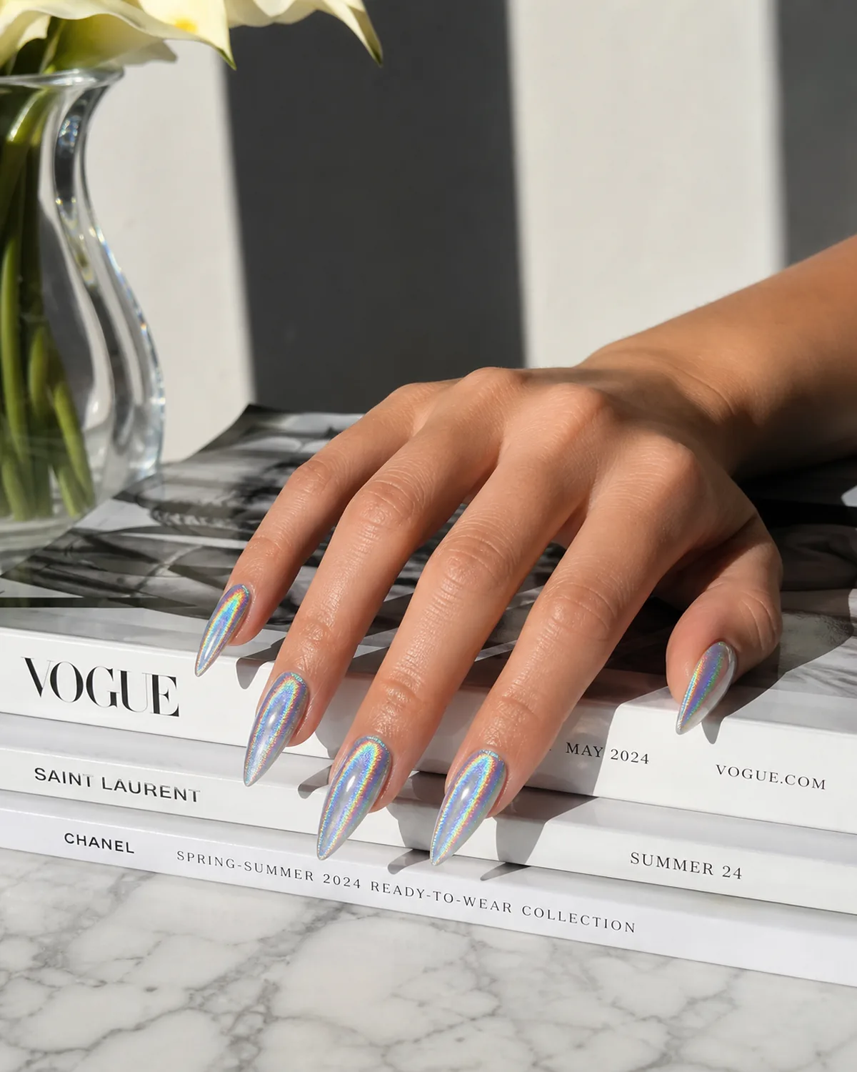 Single hand with rainbow-flash holographic silver nails on stiletto shape resting on stack of glossy white fashion magazines