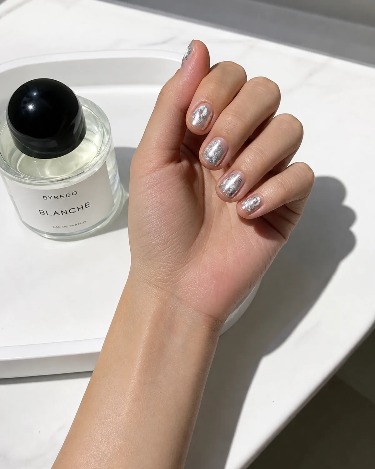 Single hand with irregularly torn silver leaf foil pieces pressed under clear gel on short round nails resting beside a glass perfume bottle on a white tray