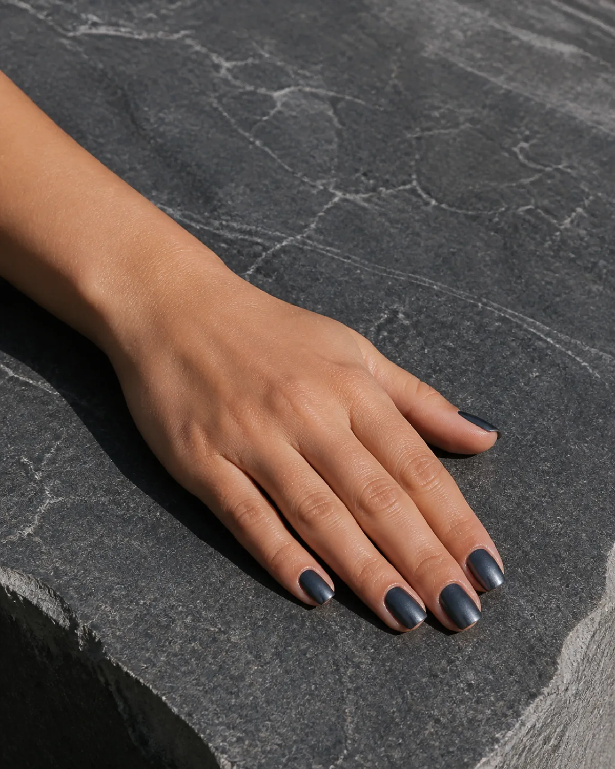 Single hand with dark charcoal-silver gunmetal satin metallic finish nails on square shape resting on dark gray stone slab