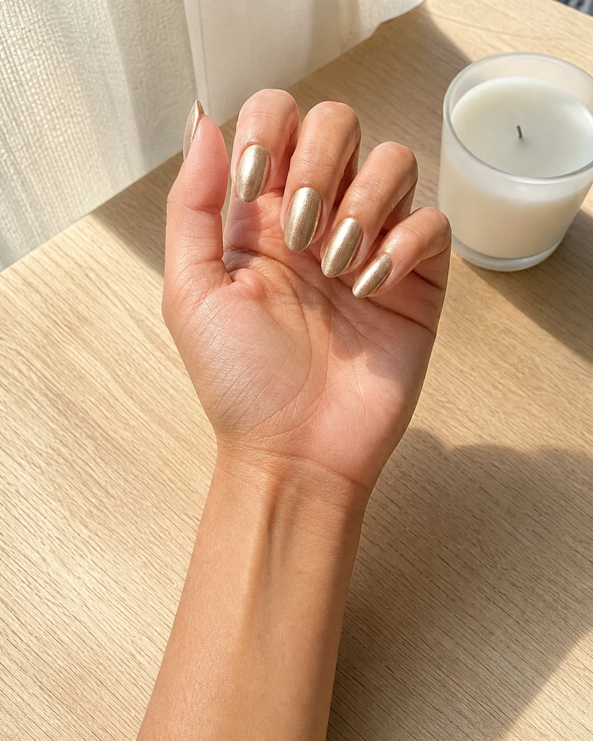 Single hand with warm-toned champagne silver metallic polish on medium almond nails resting on light wood table beside a small white candle