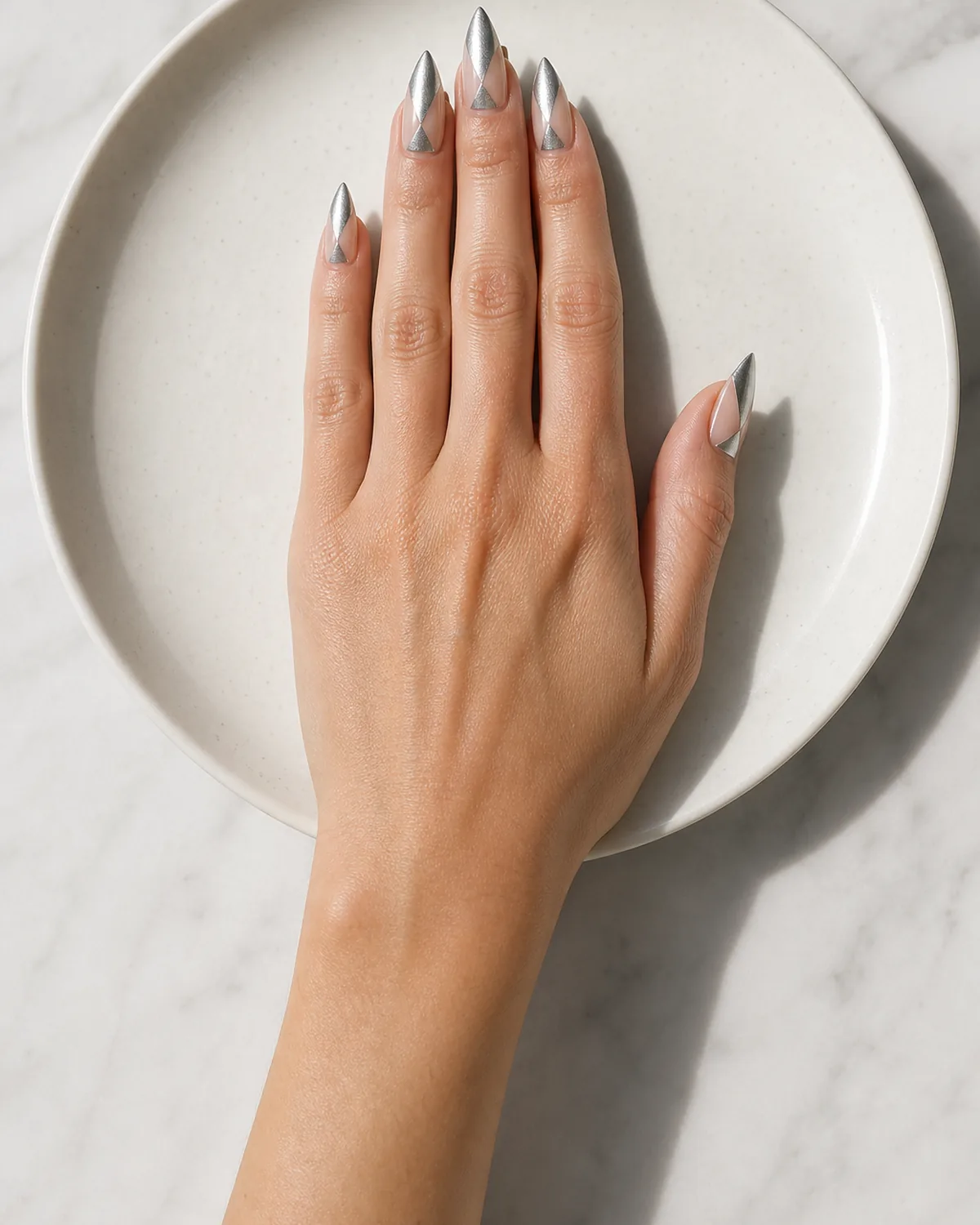 Single hand with geometric negative-space design with alternating silver metallic and bare nude triangles on medium stiletto nails resting on white ceramic plate