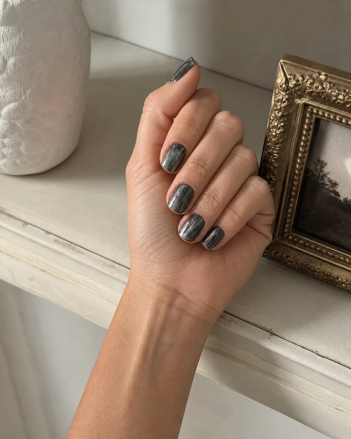 Single hand with oxidized vintage-look antique silver nails featuring matte charcoal base showing silver highlights on raised brush strokes on short squoval nails resting beside antique brass picture frame on cream shelf