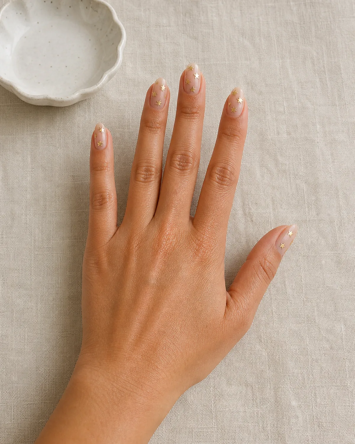 Single hand with sheer nude base and small gold metallic star stickers scattered across each nail at different angles on medium almond nails resting on cream linen tablecloth
