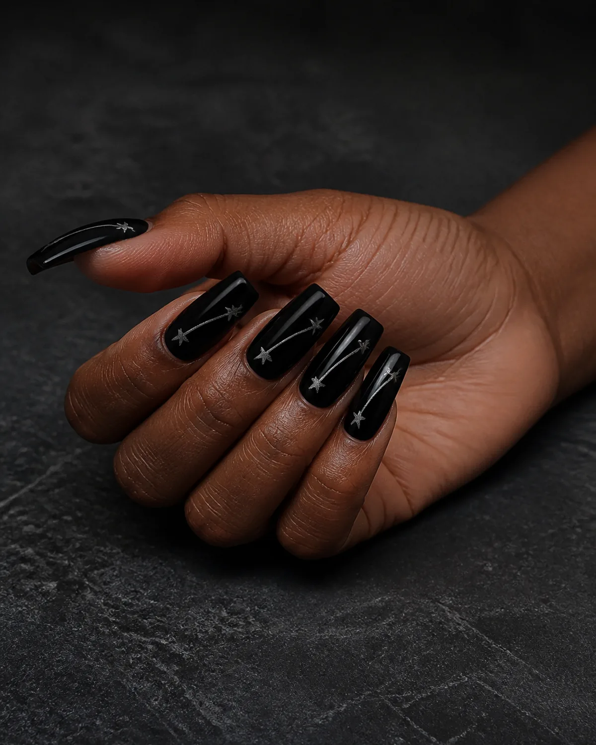 Single hand with glossy black base and thin silver shooting star trails painted diagonally across each coffin nail resting on dark charcoal slate surface