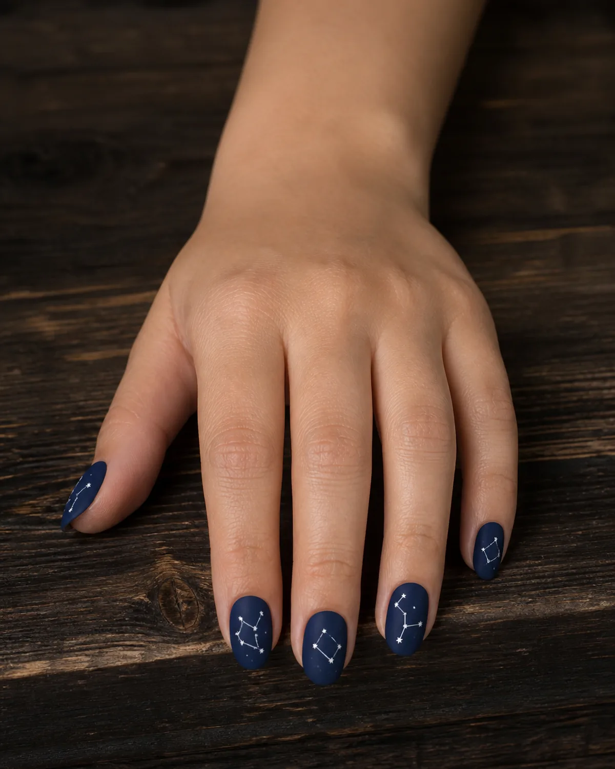 Single hand with matte dark navy blue base and tiny white dot clusters connected by fine white lines forming constellation patterns on each oval nail resting on dark wood table