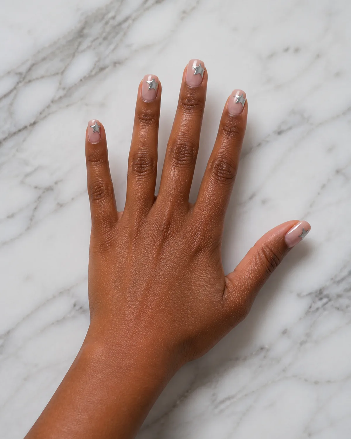 Single hand with sheer pink base and chrome silver five-pointed stars stamped at the tip of each short squoval nail resting on white marble countertop