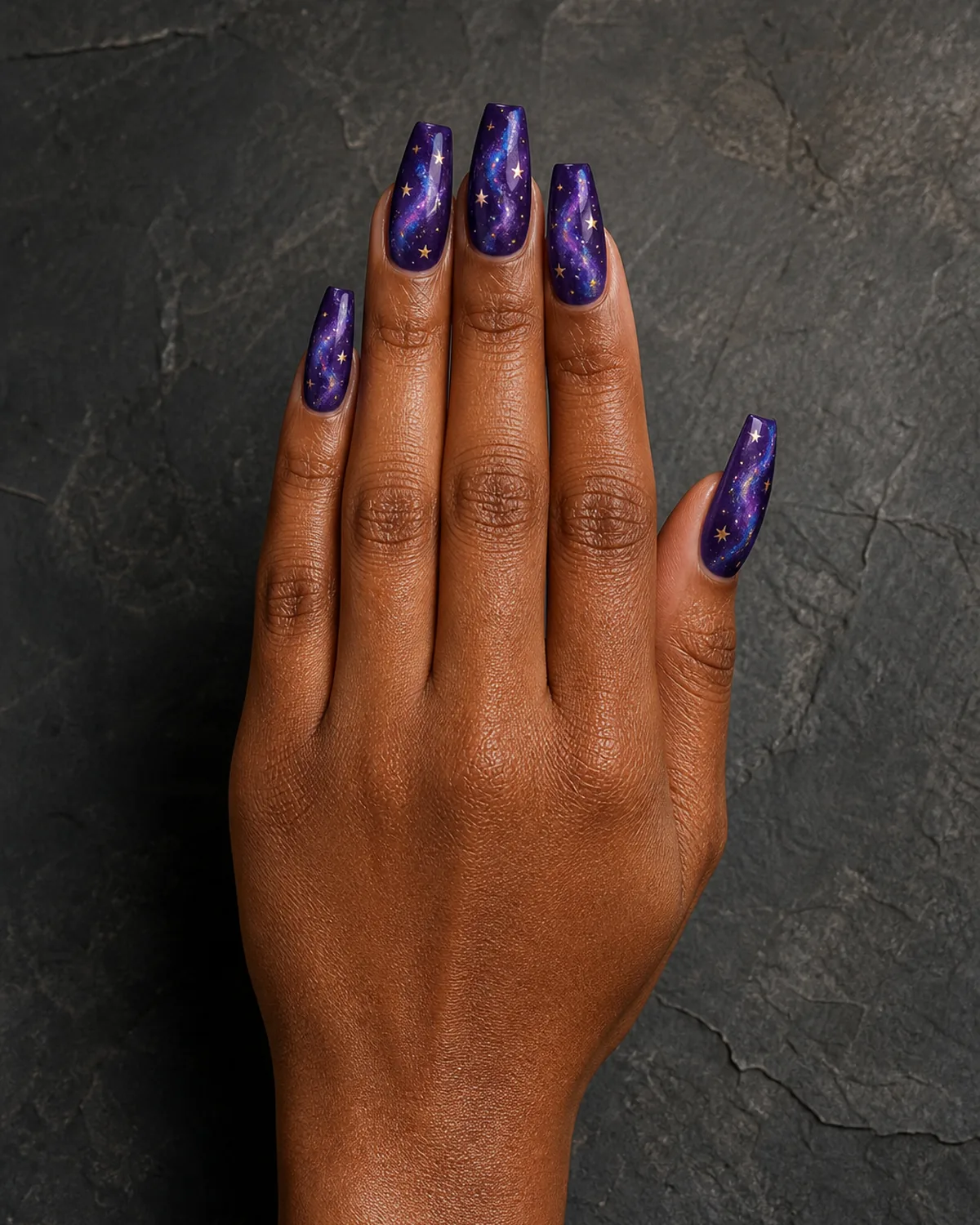 Single hand with deep purple base and swirling galaxy-like wisps of lighter purple and blue dotted with tiny gold stars on medium coffin nails resting on dark gray stone surface