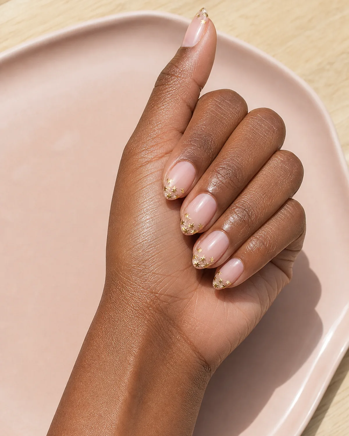 Single hand with sheer pink base and tip area of each oval nail filled with cluster of small gold metallic stars instead of a solid French tip resting on blush-pink ceramic tray