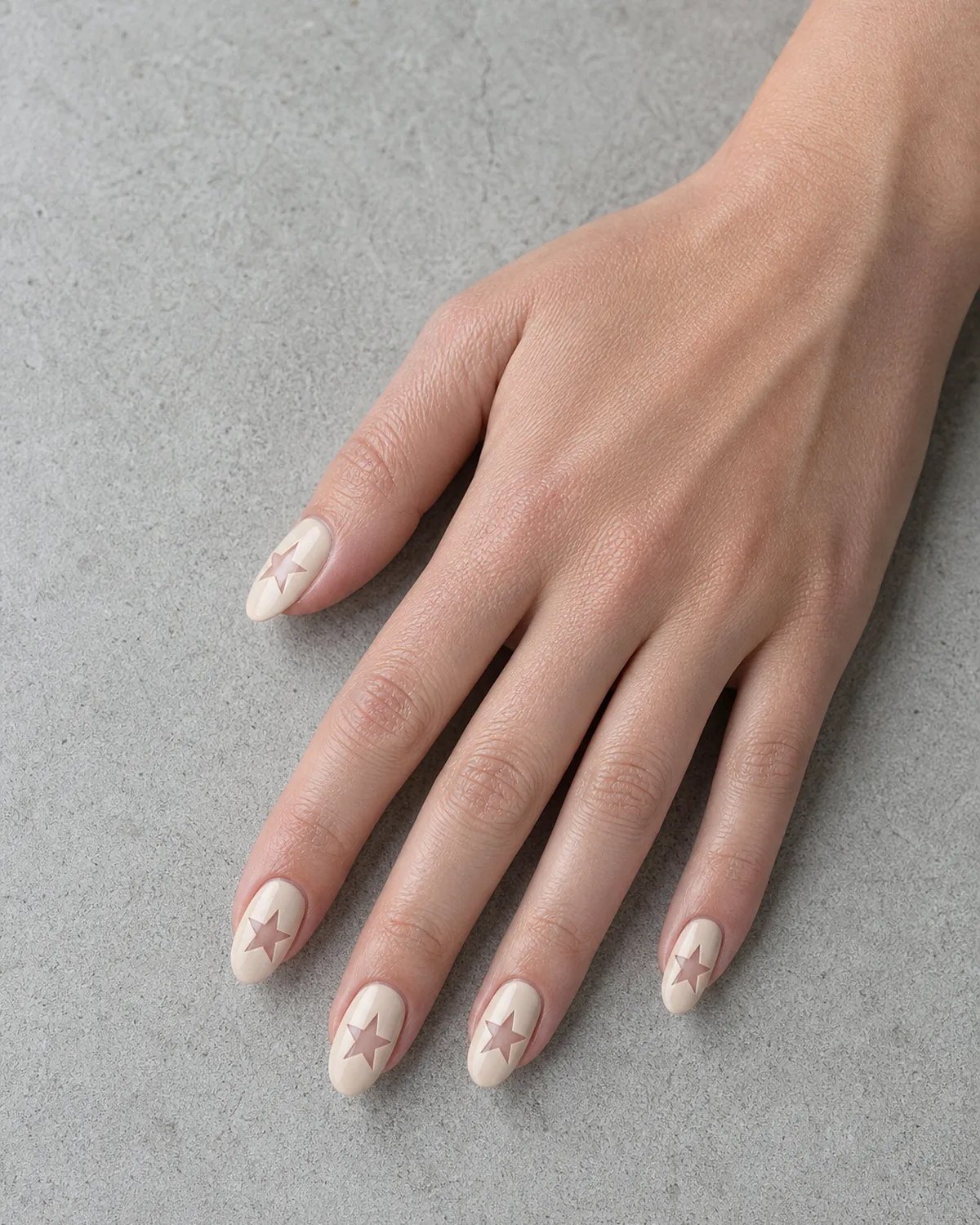 Single hand with clear bare nail base and opaque soft beige sections creating a star cutout negative space pattern on medium almond nails resting on pale gray concrete surface