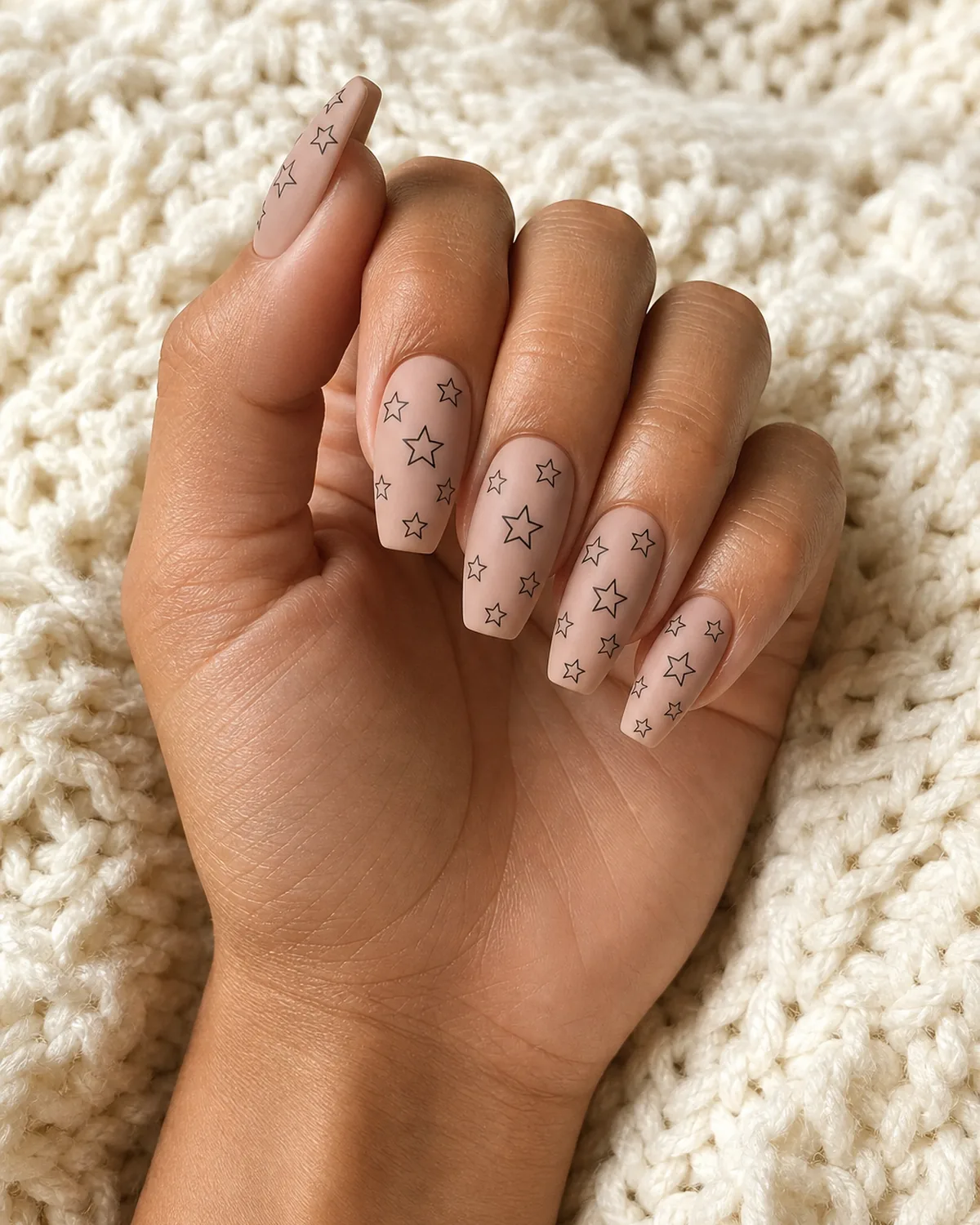 Single hand with matte nude base and thin black outlined five-pointed stars in varying sizes scattered across each medium coffin nail resting on warm cream knit blanket