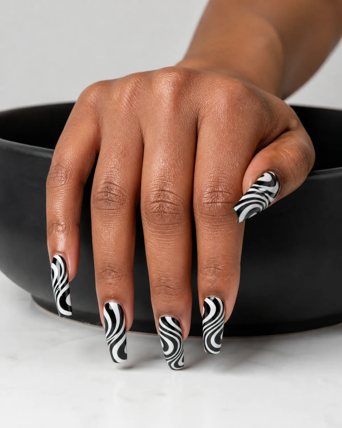 Single hand with bold alternating black and white retro swirl lines in 1960s psychedelic pattern on long coffin shaped nails draped over edge of matte black ceramic bowl