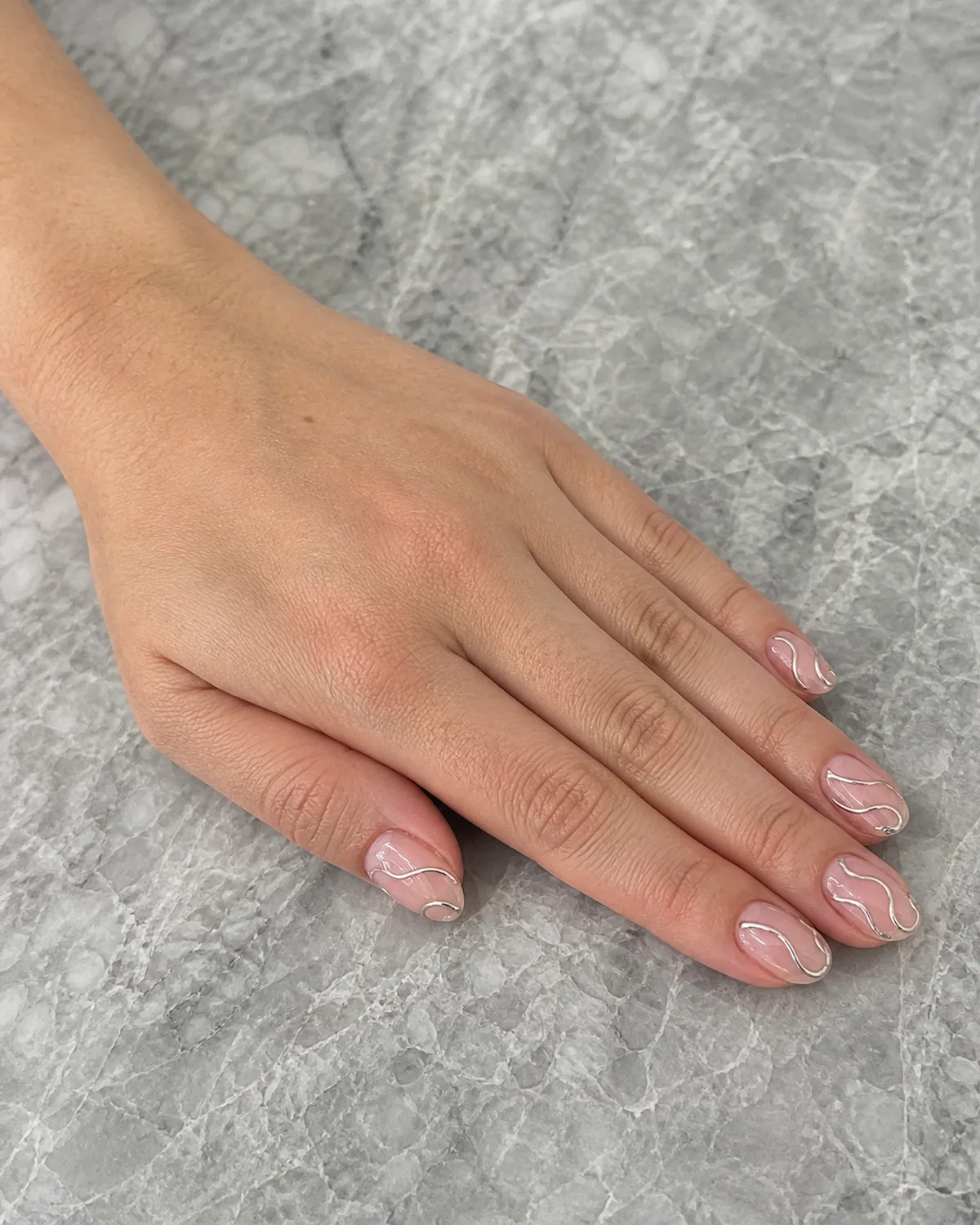 Single hand with thin raised chrome silver swirl lines over sheer pink jelly base nail art on short almond shaped nails resting flat on gray marble countertop