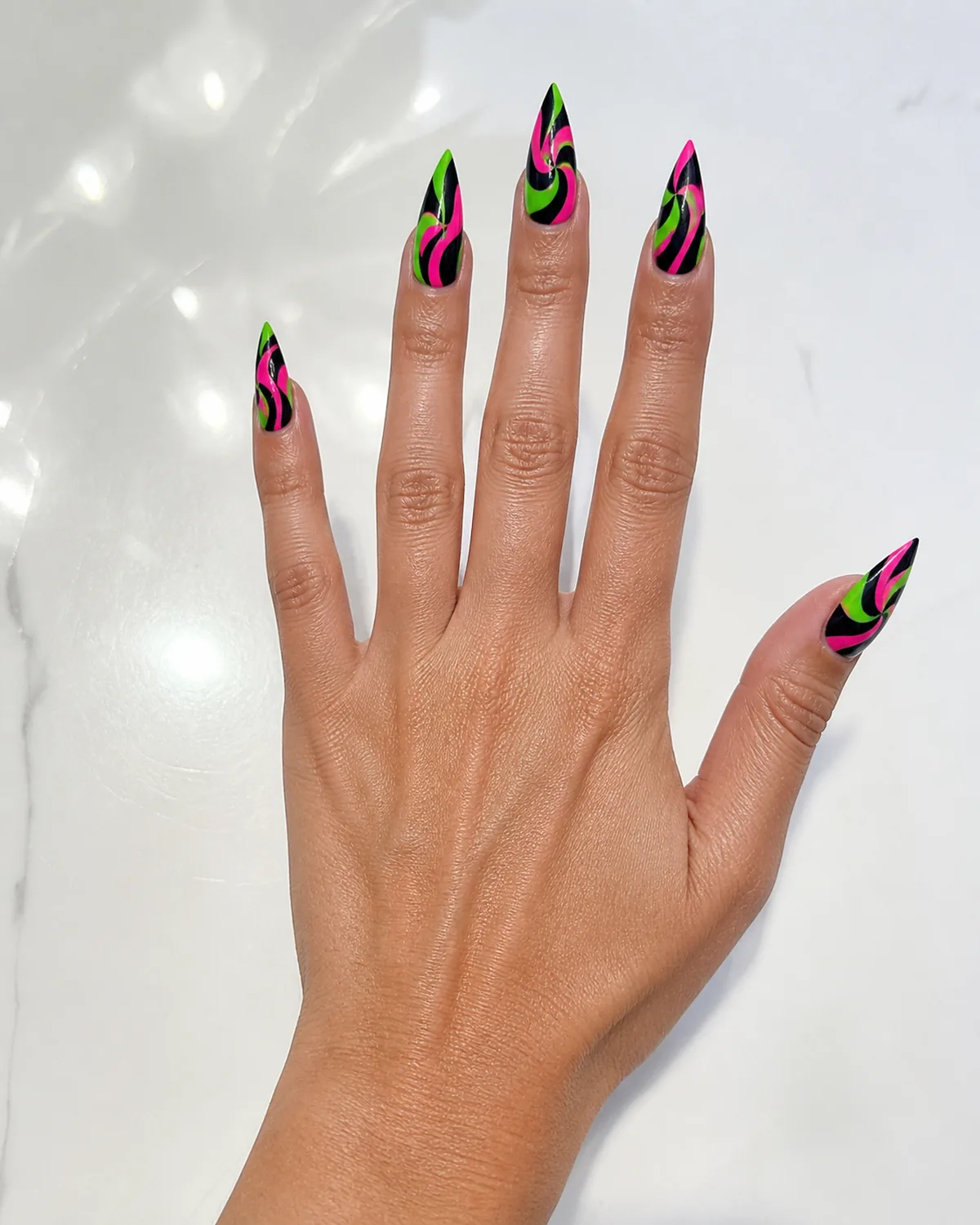 Single hand with electric neon green and hot pink swirl nail art in bold high-contrast curves over black base on long stiletto shaped nails resting on glossy white acrylic surface