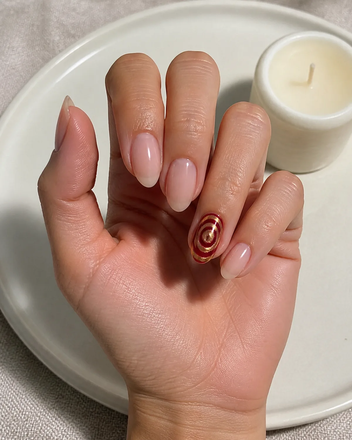 Single hand with abstract concentric swirl circles in deep burgundy and gold on ring finger accent nail with remaining nails in plain sheer nude on medium oval shaped nails lying on white ceramic plate beside small white candle
