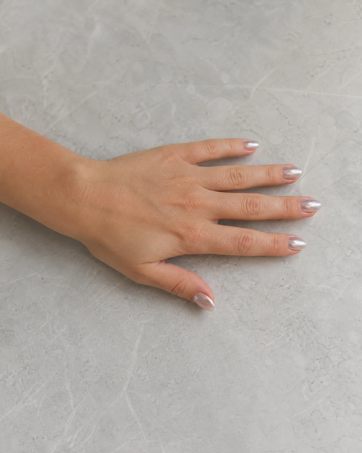 Single left hand with clear chrome glazed nails on short almond shape palm down on light gray stone surface