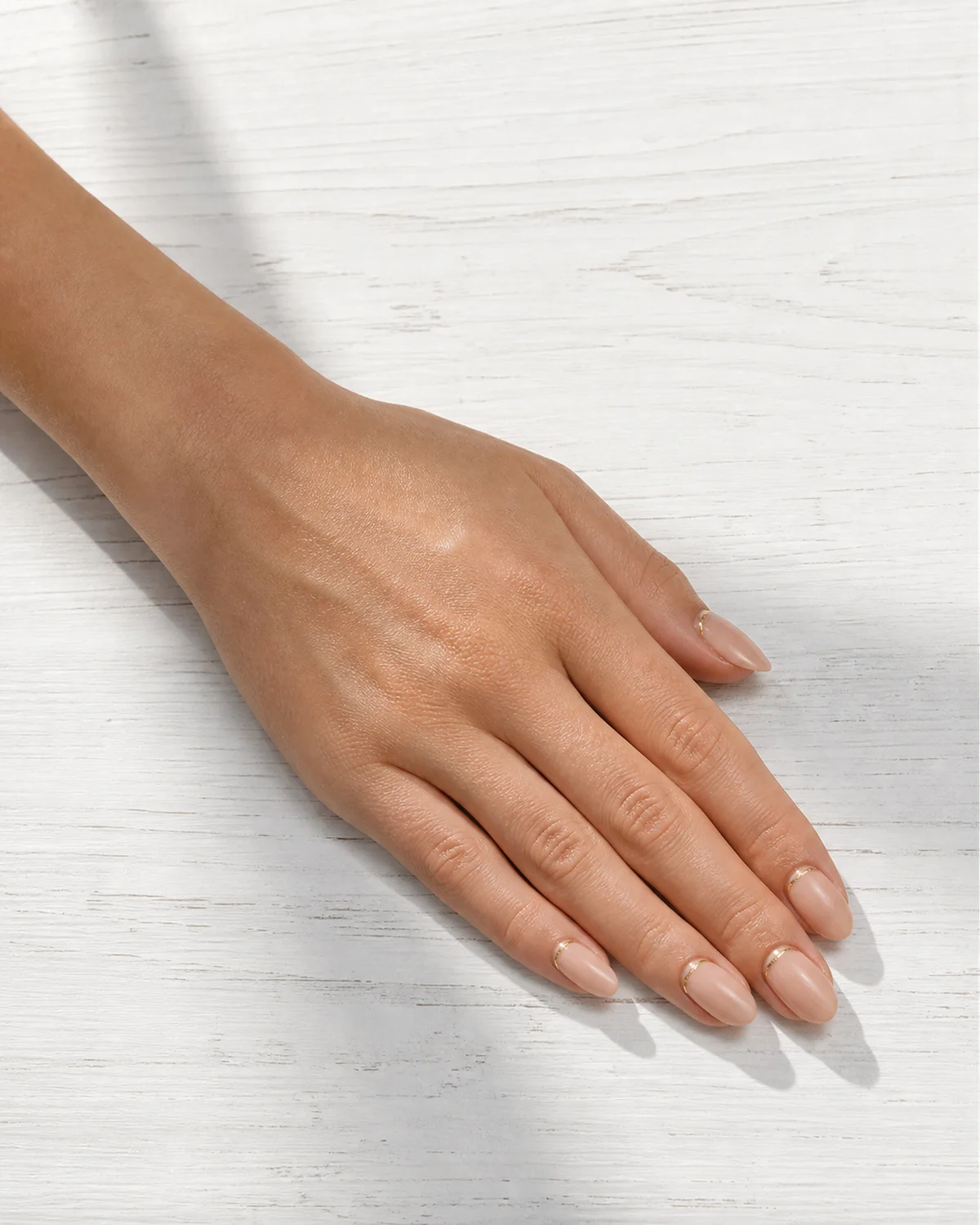 Single left hand with warm nude almond nails and thin gold foil line at the cuticle palm down on white wood table