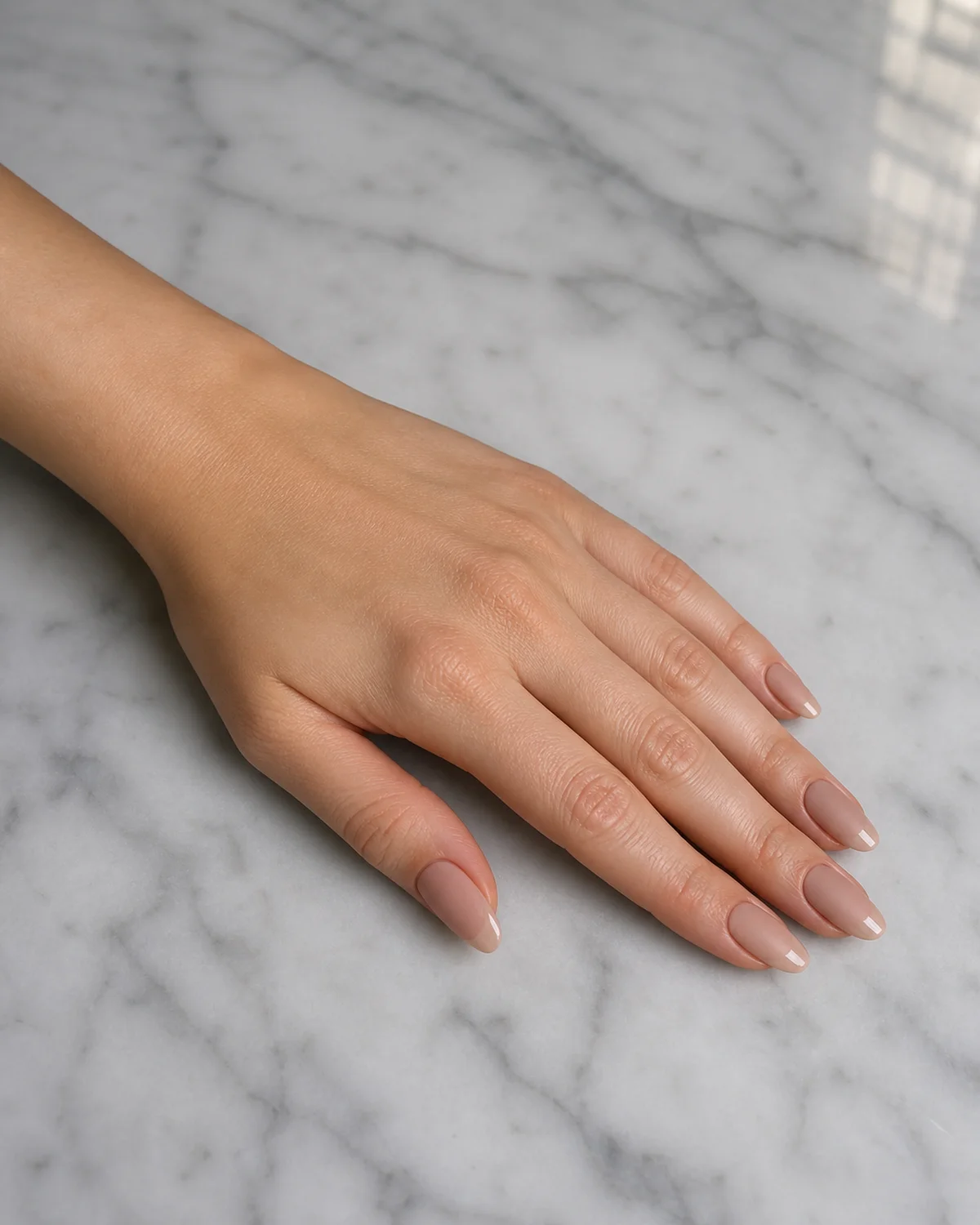Single left hand with matte warm nude nails and glossy tip on medium almond shape palm down on pale gray marble counter