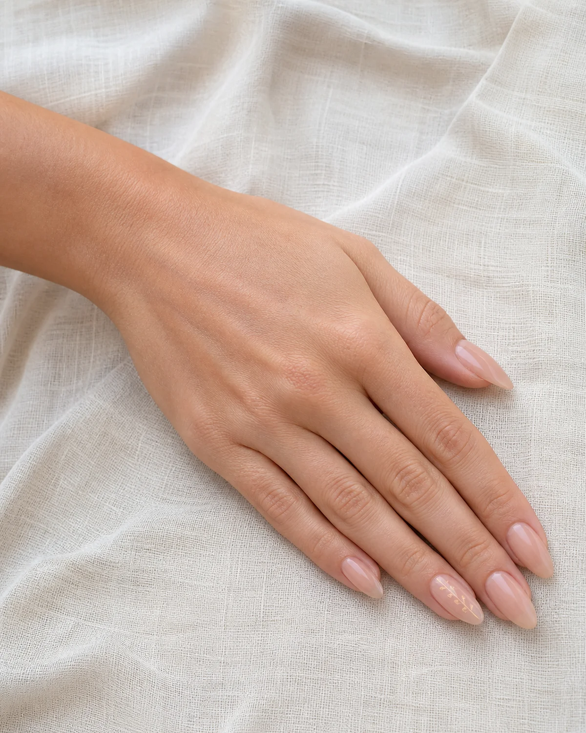 Single left hand with sheer nude almond nails and a soft gold vine on the ring finger palm down on off-white textured linen