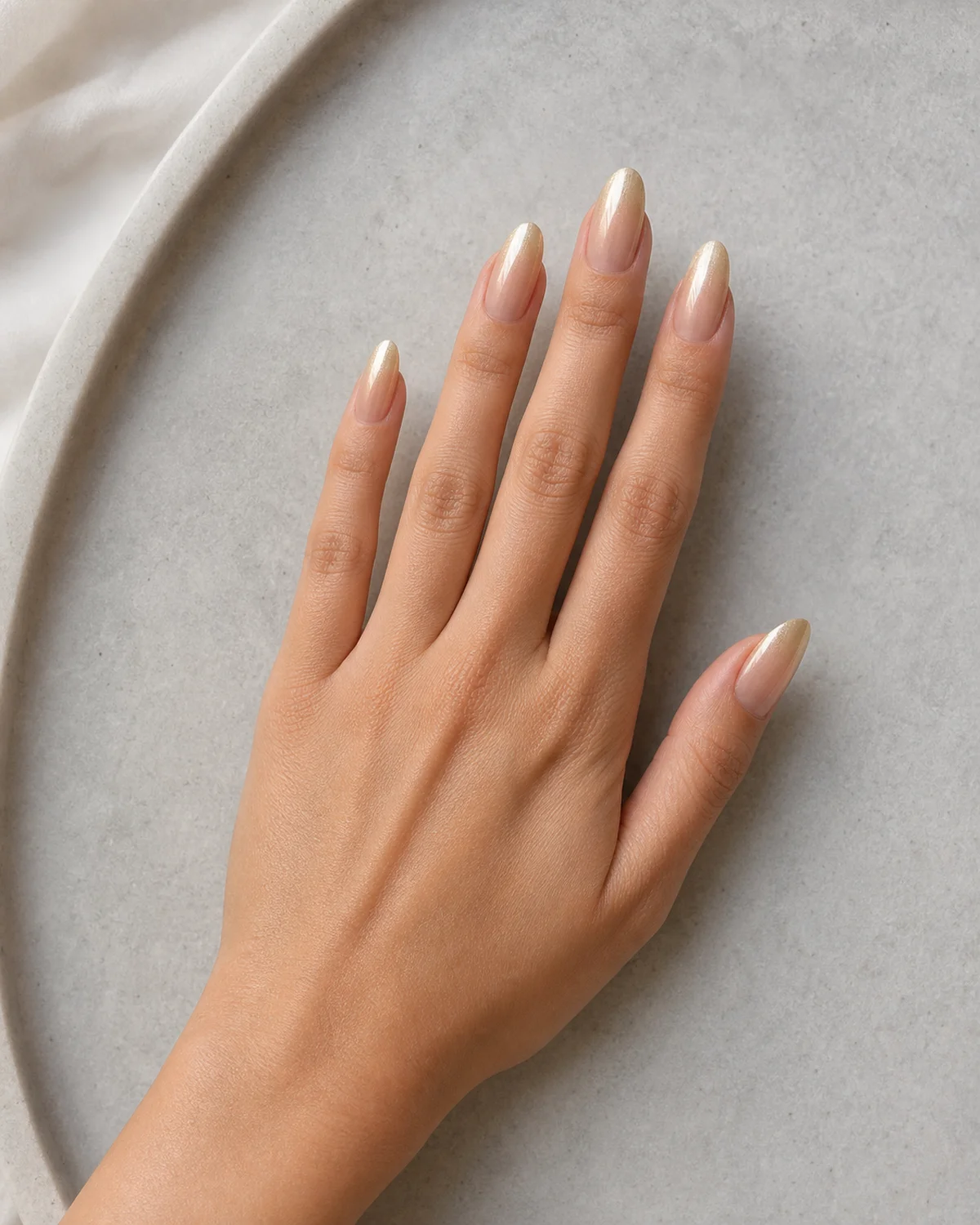 Single left hand with nude-to-champagne gold vertical gradient nails on medium almond shape palm down on pale gray stone tray
