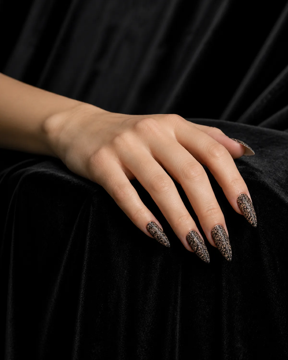 Single left hand resting on dark velvet fabric with long stiletto nails featuring black lace pattern over warm nude base
