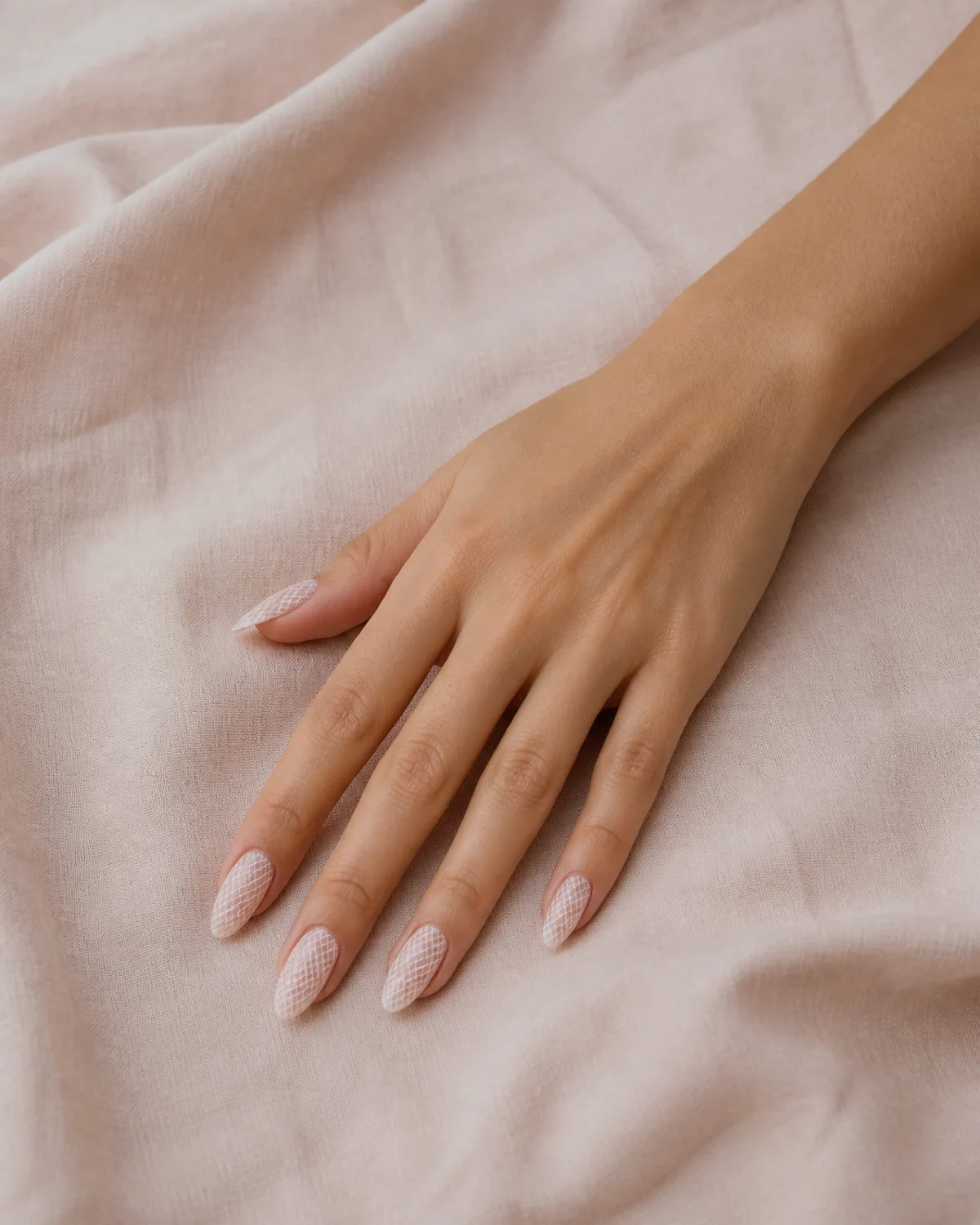 Single right hand palm down on blush-toned linen with long almond nails featuring fine mesh-style lace grid pattern in barely-there white over warm nude gel base