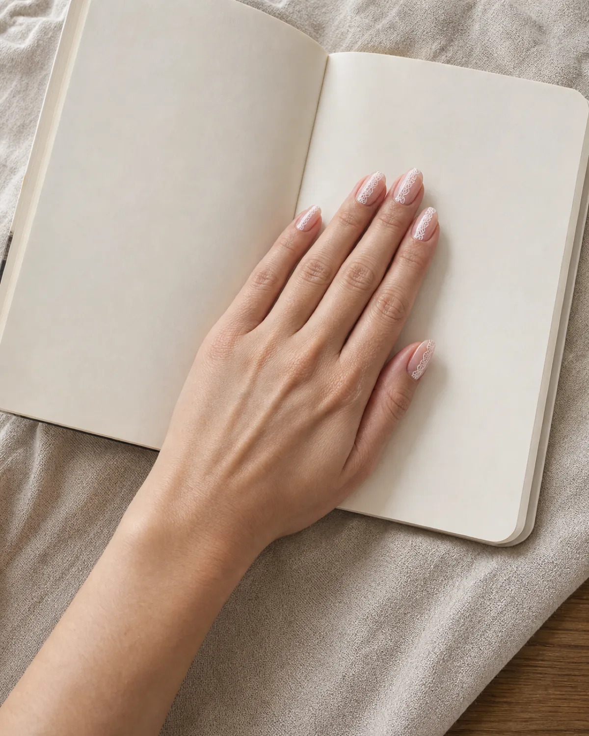 Single left hand resting on open blank journal with medium oval nails featuring single vertical lace stripe from cuticle to free edge down center of each nail on sheer pink base