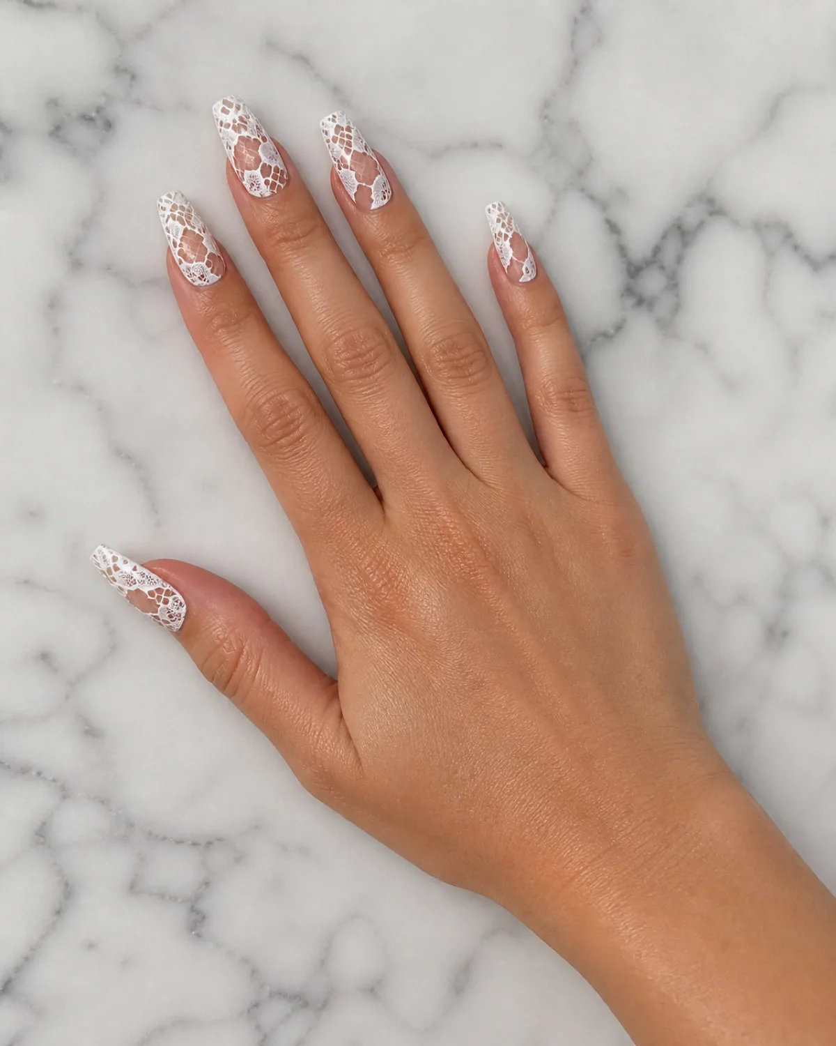 Single right hand palm down on white marble surface with long coffin nails featuring white lace pattern painted in opaque white with sections of bare natural nail exposed as negative space