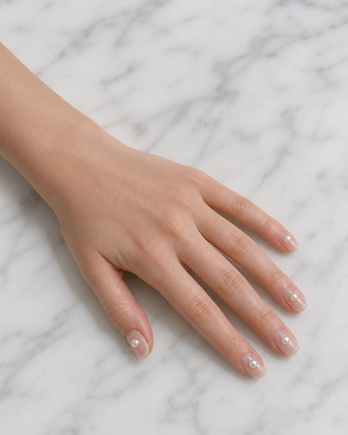 Single left hand with medium oval nails each bearing one small faux pearl bead on clear gel base palm down on white marble surface