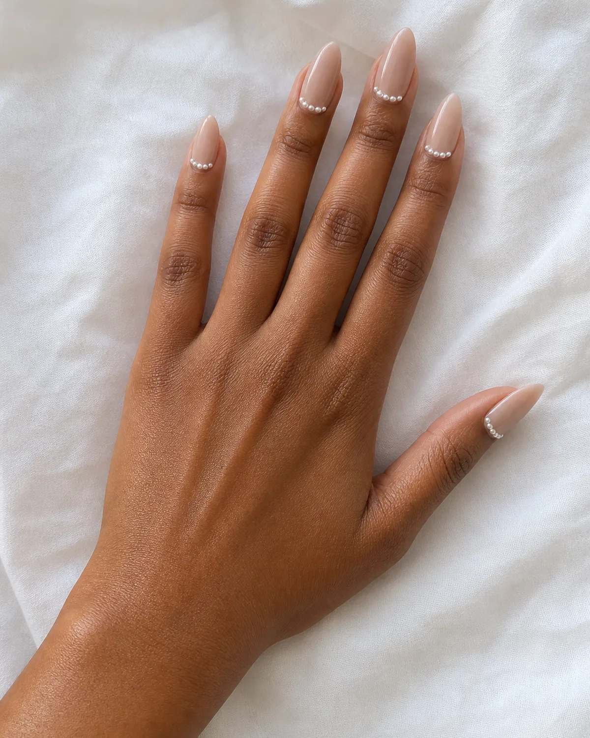 Single left hand with long almond nails and tiny pearl cuff along the cuticle line palm down on white cotton fabric