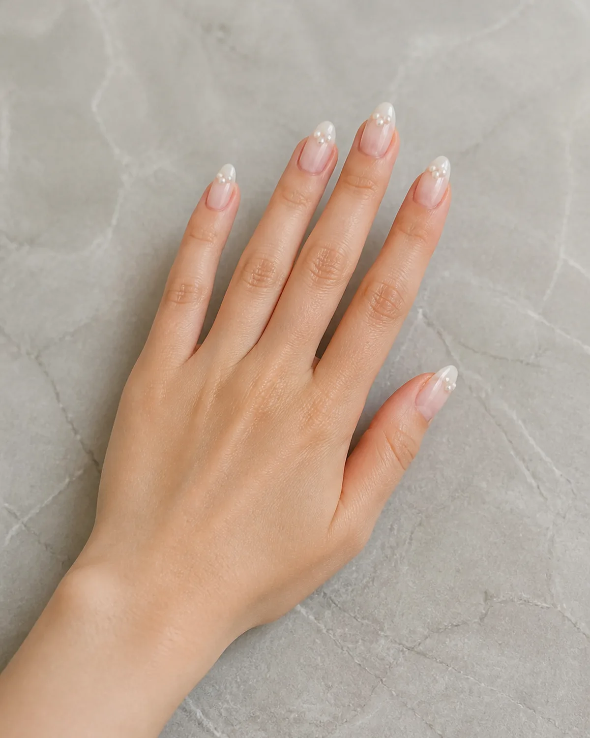 Single left hand with milky white jelly base and pearl cluster at the tip of medium almond nails palm down on pale gray stone surface