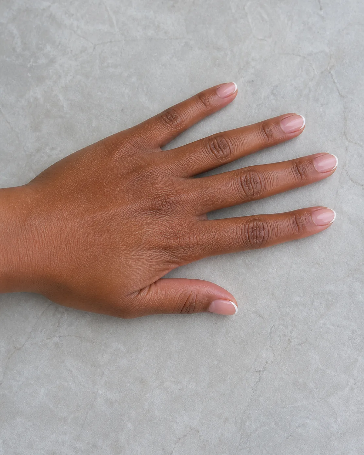 Single left hand with micro French thin white tip line on short almond nails palm down on pale gray stone surface