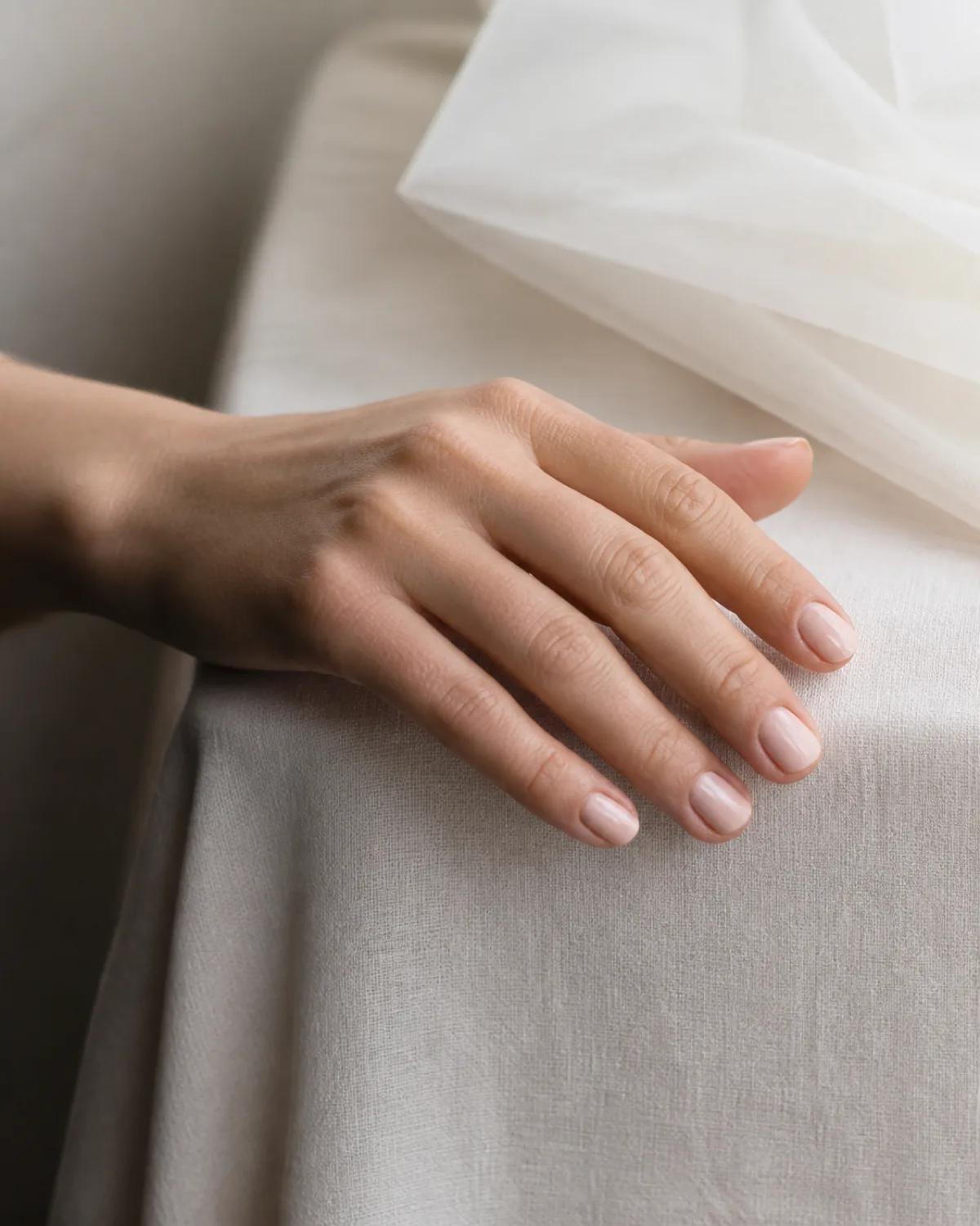 Single hand with sheer milky polish on short natural nails draped over linen-covered table edge