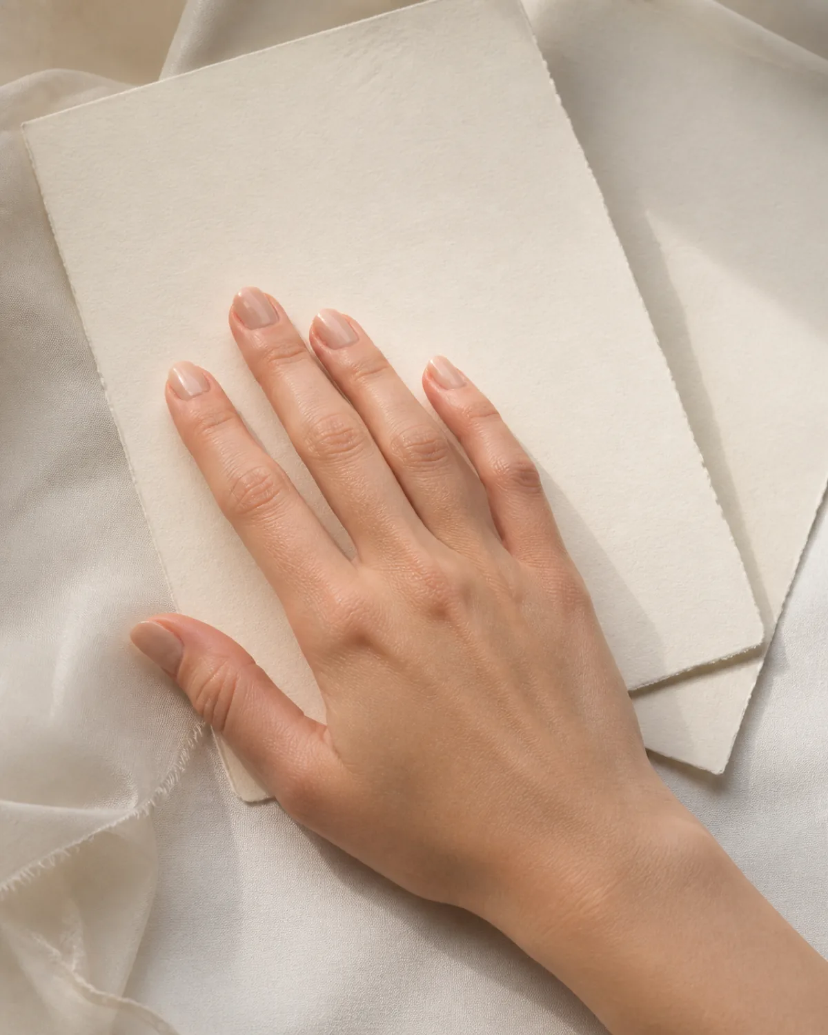 Single hand with barely-there beige polish on round nails resting on cream wedding invitation paper