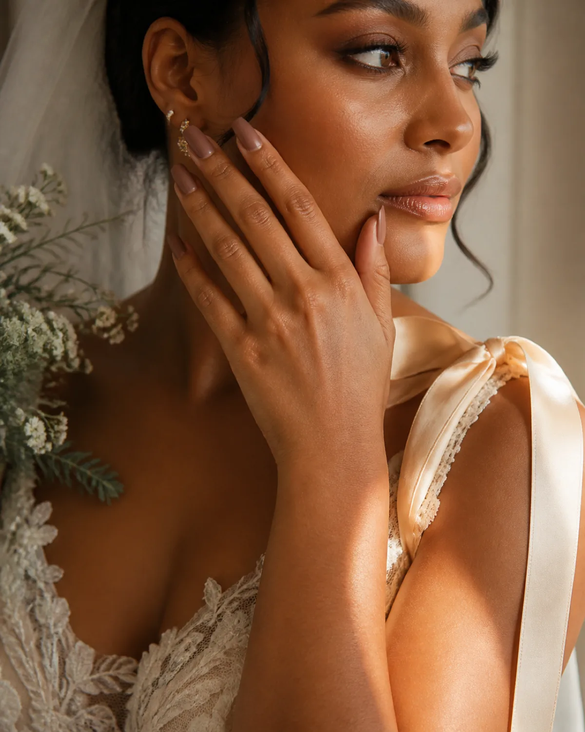 Single hand near cheek in beauty editorial pose showing warm nude matte coffin nails on deeper skin tone