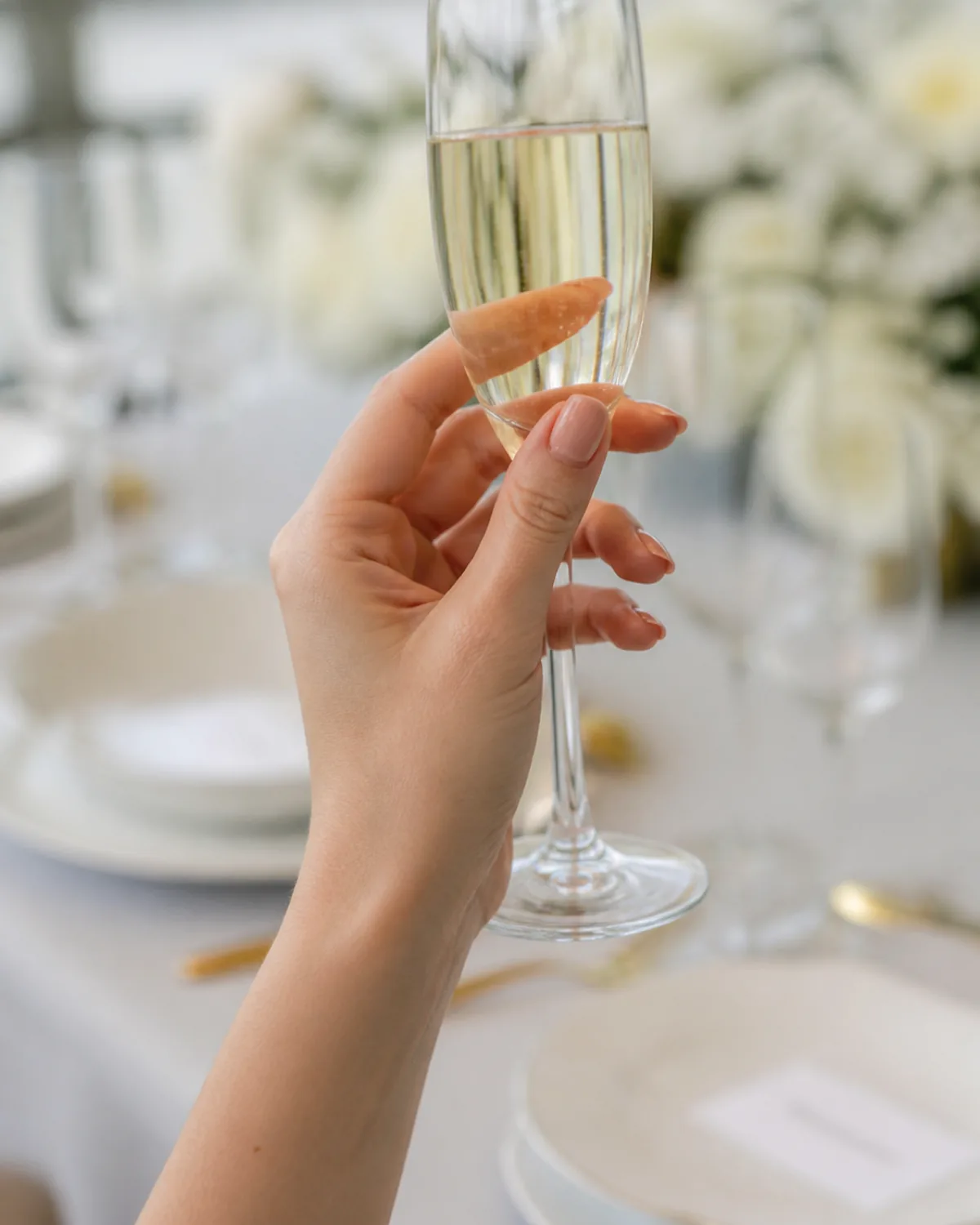Single hand holding champagne flute by stem showing rose water tinted sheer polish on short almond nails