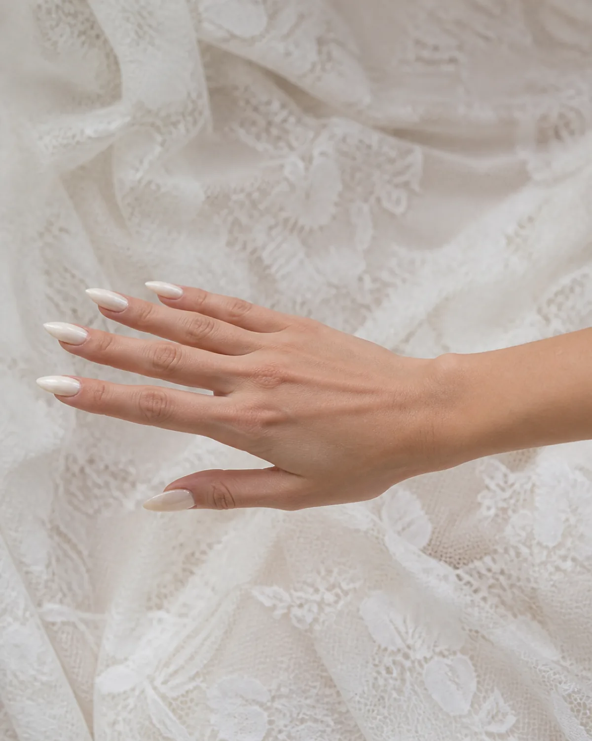 Side profile of single hand with off-white glossy stiletto nails extended against white lace background