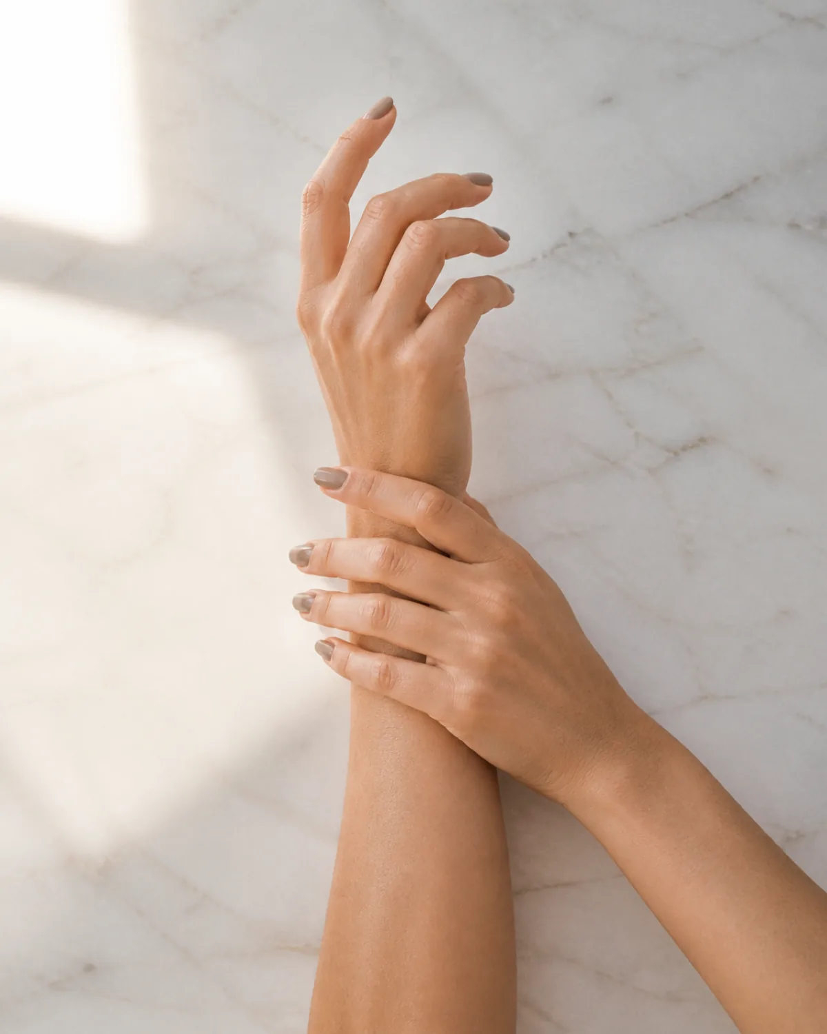 Two hands with one holding the opposite wrist showing greige polish on squoval nails against marble