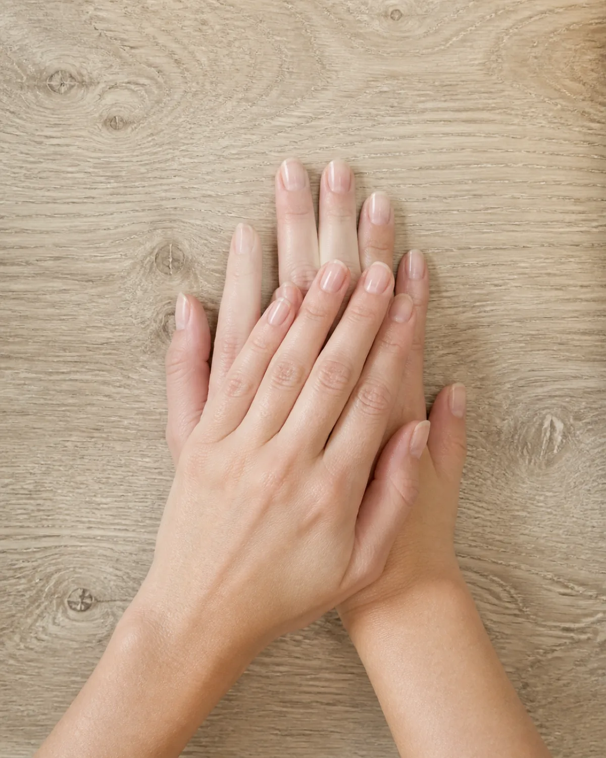 Two hands stacked on light oak wooden surface showing bare buffed nails with clear topcoat and natural shine