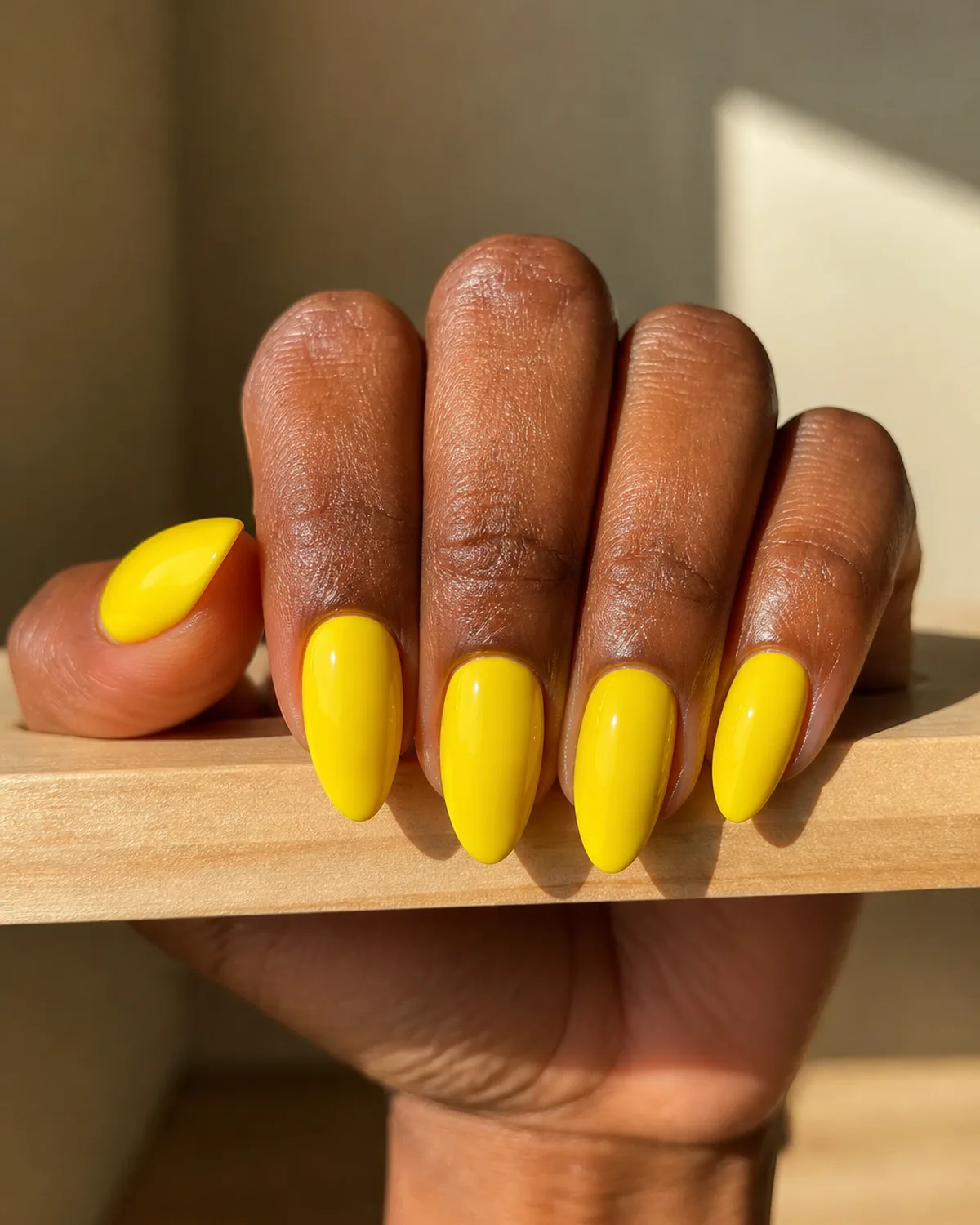 Single hand with bright true-lemon yellow glossy polish on medium almond nails with fingers curled over the edge of a light wooden shelf