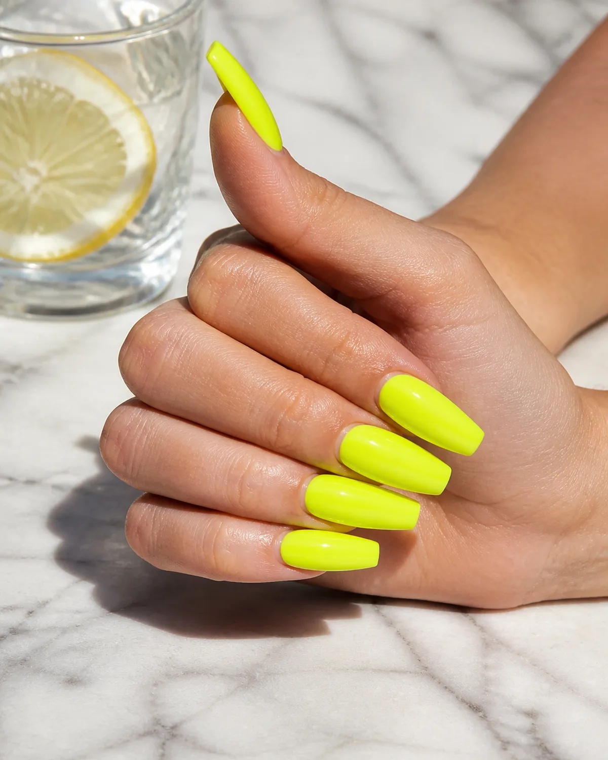 Single hand with electric neon highlighter-yellow polish on long coffin nails resting on its side on white marble beside a glass of lemon water