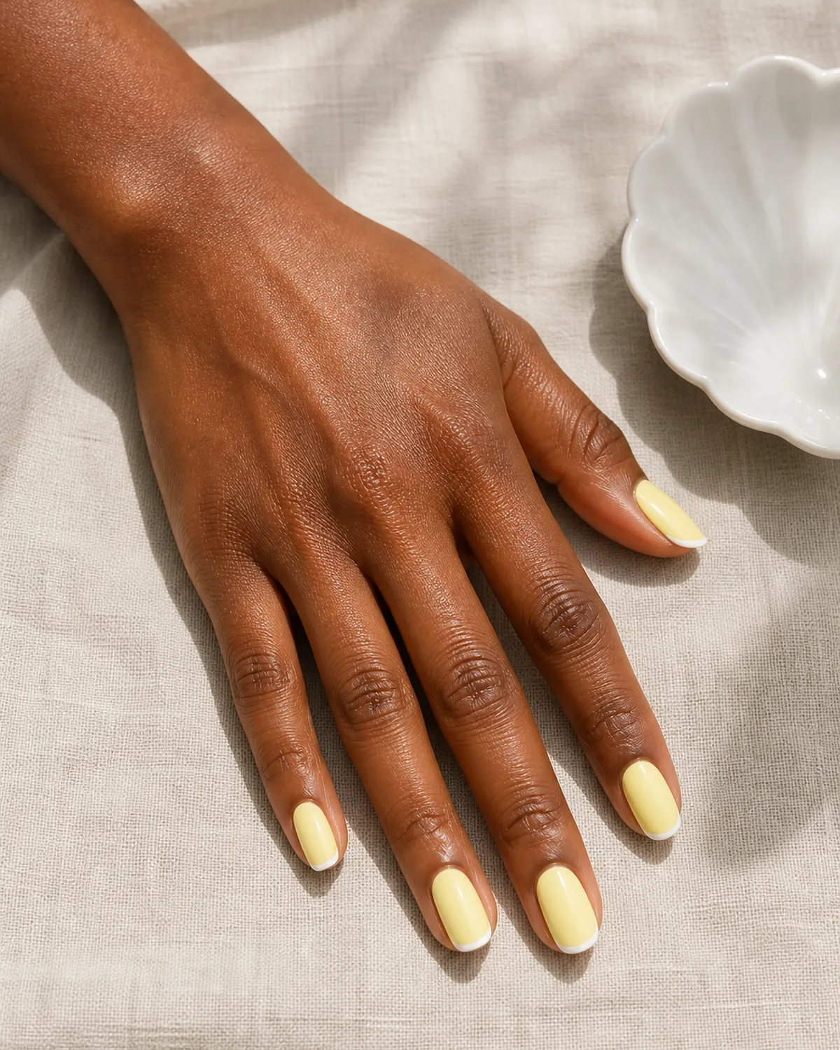 Single hand with sheer pastel baby yellow base and white micro French tips on oval nails resting palm down beside a white porcelain dish on linen